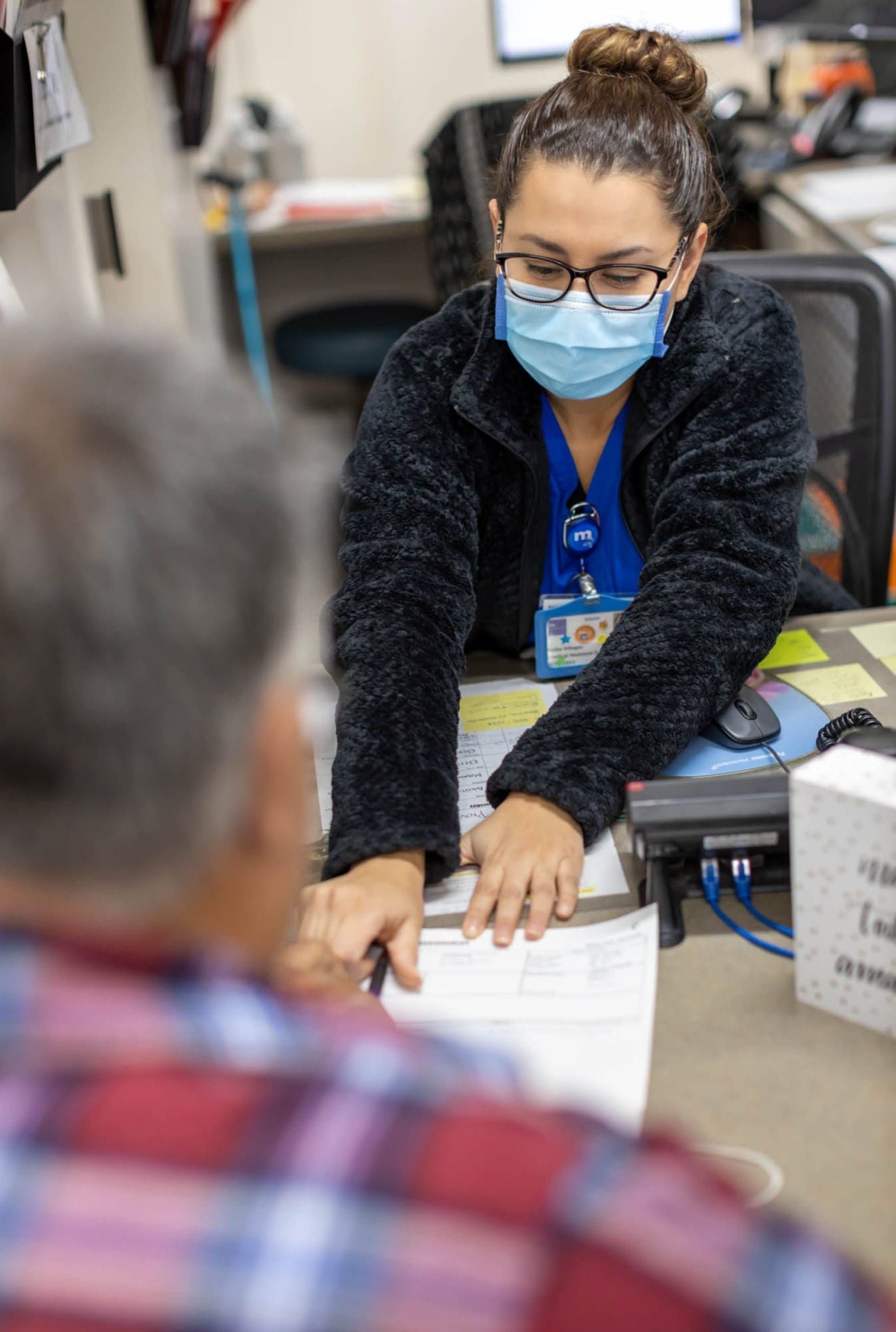 A woman wearing glasses and a face mask sits at a desk, pointing to a document in front of a man in a plaid shirt, likely explaining or reviewing paperwork with him in an office setting.