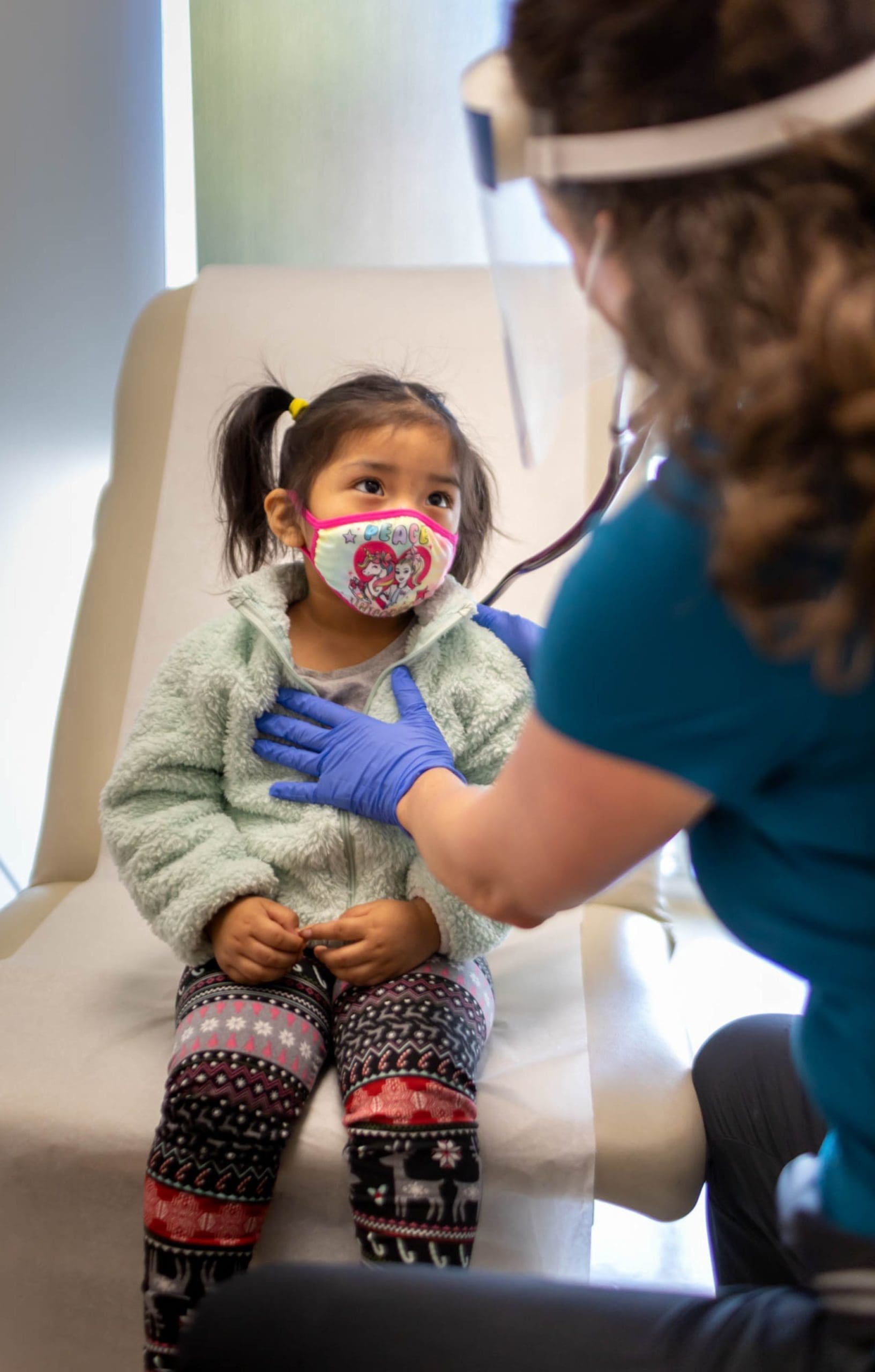 A young child wearing a colorful mask sits on a chair while a healthcare worker in gloves and a face shield gently places a hand on the childs chest during a medical checkup.