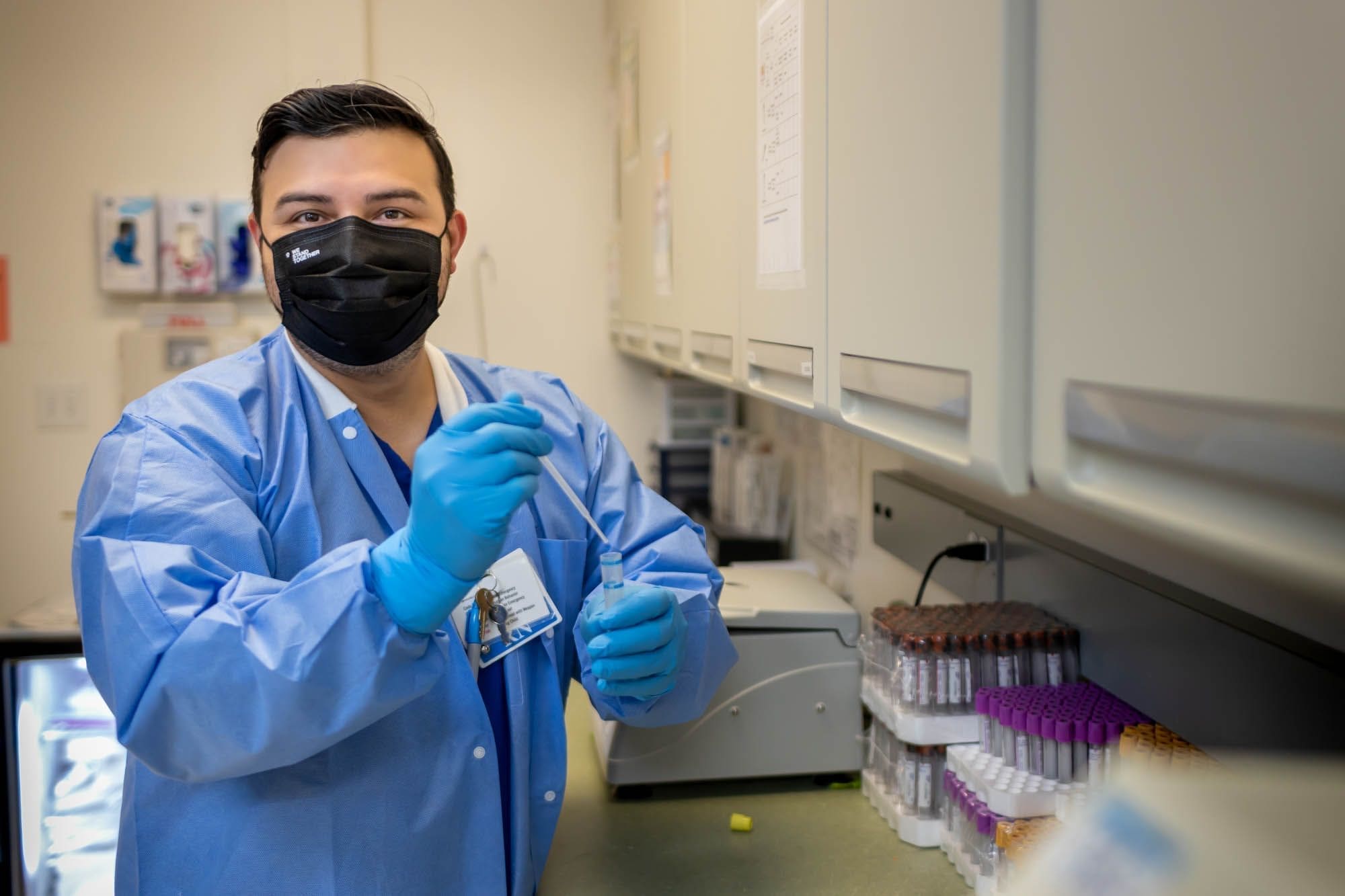 A lab technician wearing a black face mask, blue gloves, and a blue lab coat holds a pipette in a laboratory, with test tubes and equipment on the counter beside him.
