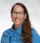 A woman with long brown hair and glasses smiles at the camera. She is wearing a blue collared shirt and earrings, with a white and gray background.