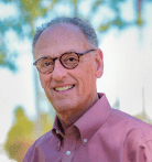 Smiling older man with short gray hair and glasses wearing a mauve button-down shirt, standing outdoors with blurred green trees in the background.