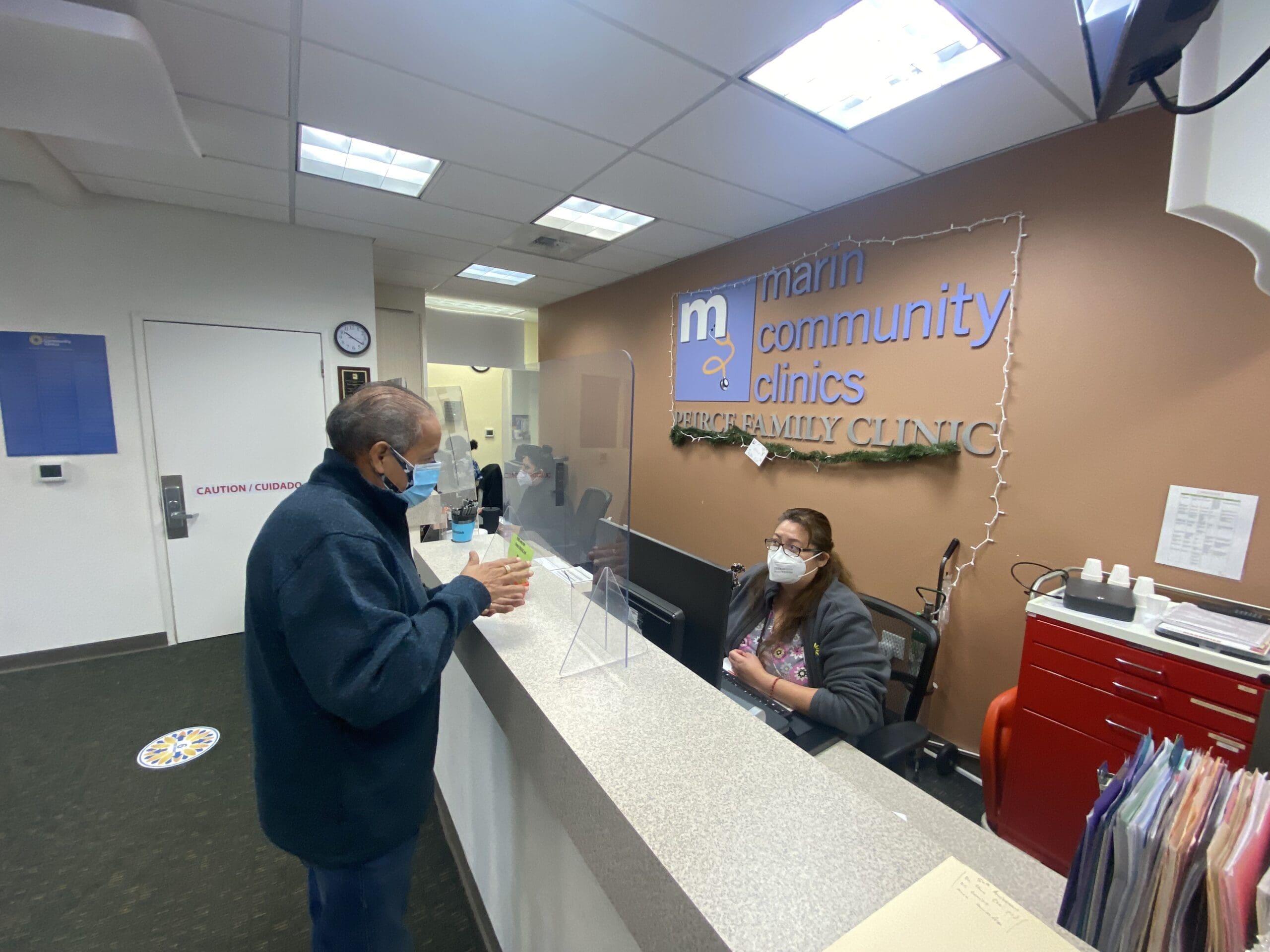 An elderly man wearing a mask speaks to a masked receptionist behind a transparent barrier at the front desk of Marin Community Clinics. Office signs, files, and safety measures are visible in the clinic lobby.