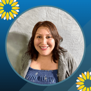 A woman with medium-length brown hair smiles at the camera. She is wearing a blue patterned top and a gray cardigan. The image has a blue border with yellow and white flower graphics in two corners.