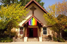 A church building with a rainbow pride flag hanging above the entrance, surrounded by trees and bright sunlight.