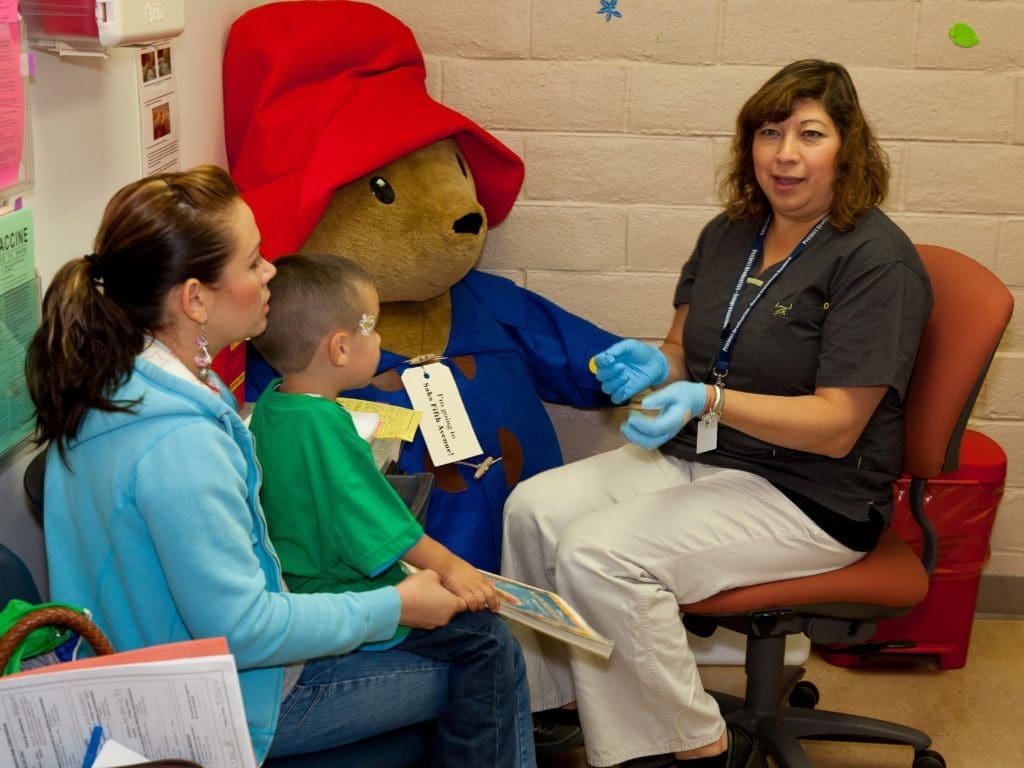 A woman in a blue hoodie sits with a young boy on her lap in a clinic, talking to a healthcare worker in gloves. Behind them is a large Paddington Bear stuffed toy dressed in a coat and hat.