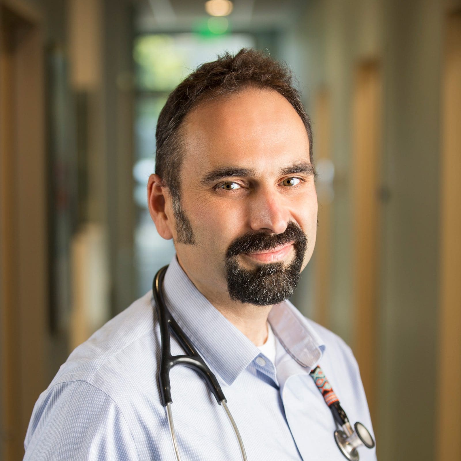 A man with dark hair and a beard, wearing a light blue shirt and a stethoscope around his neck, smiles slightly while standing in a brightly lit hallway.