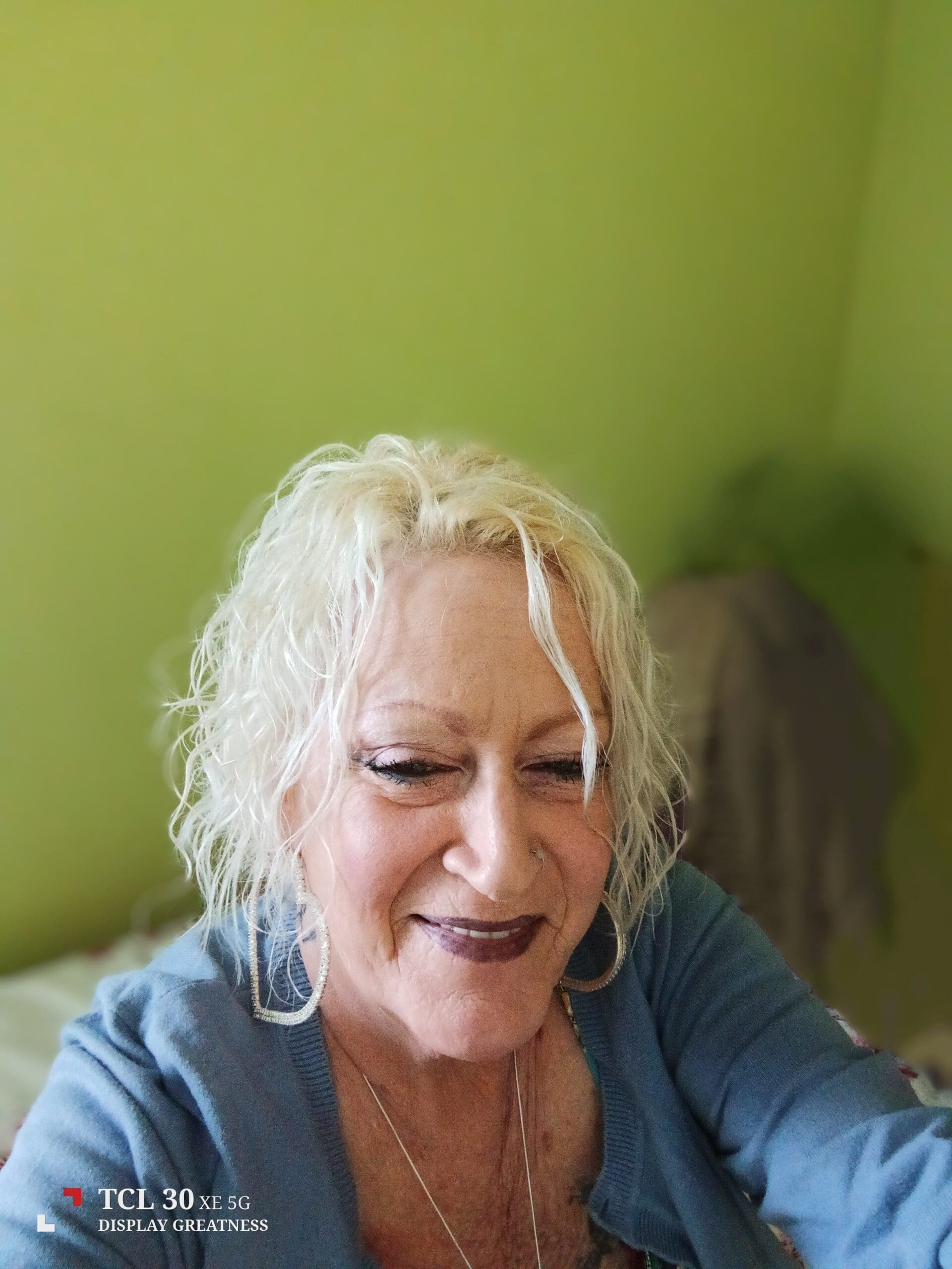 A smiling older woman with short, curly, blonde hair is sitting indoors against a light green wall. She is wearing large hoop earrings, a blue top, and dark lipstick. The photo is slightly out of focus in the background.