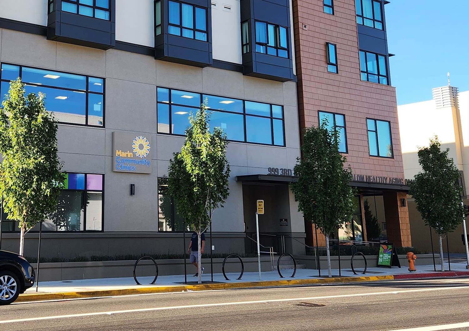 A modern building with “Marin Community Clinics” sign, large windows, young street trees, a sidewalk, a bike rack, and someone walking past. There is a car parked on the street in front.