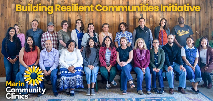 A diverse group of people pose together indoors, smiling, under a sign reading Building Resilient Communities Initiative. The Marin Community Clinics logo is visible in the lower left corner.