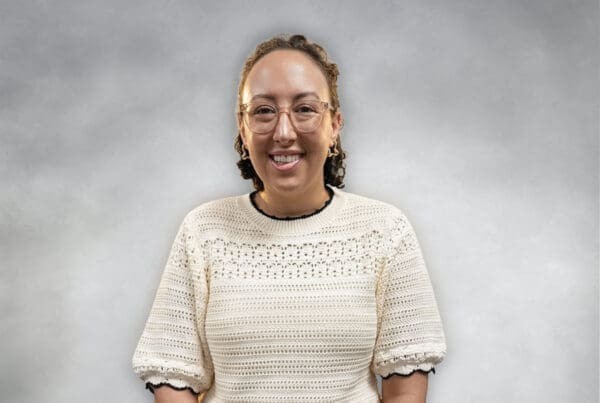 A smiling woman wearing glasses and a cream-colored crochet top against a gray background.