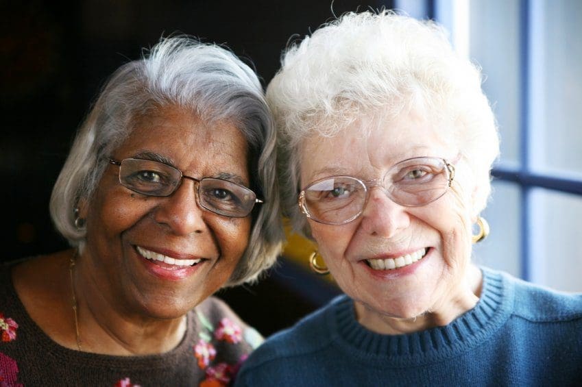 Two older women smiling and posing closely together, both wearing glasses and sweaters, with natural light coming from a window beside them.