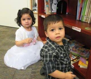 Two young children, a girl in a white dress and a boy in a plaid shirt, are sitting on the floor in front of a bookshelf filled with books. Both are looking towards the camera.