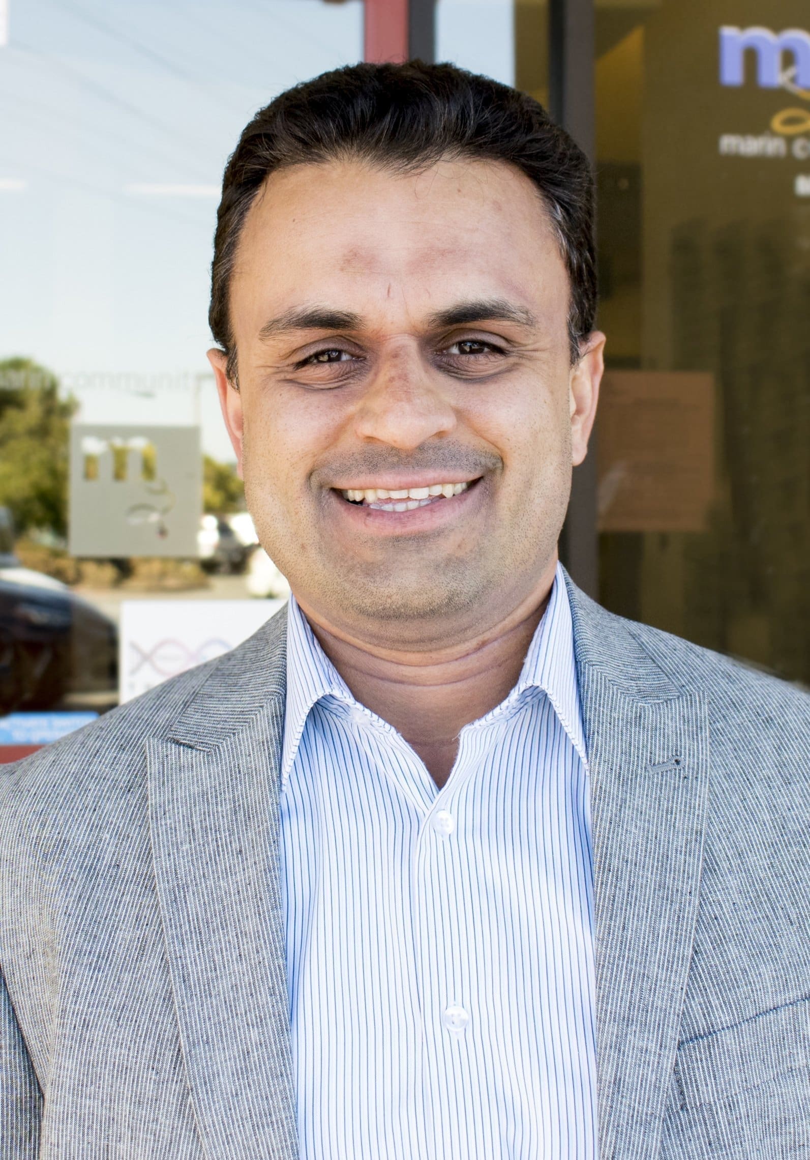 A man with short dark hair wearing a light gray blazer and a blue striped shirt smiles while standing outside a building with glass doors.