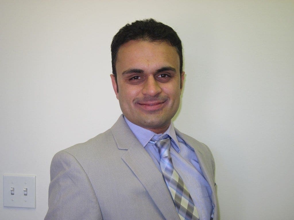 A man in a light gray suit and striped tie stands in front of a plain white wall, smiling at the camera. A light switch and electrical outlet are visible on the left side of the image.