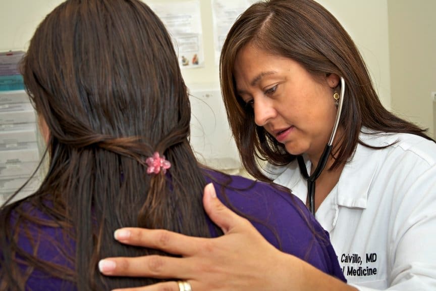 A doctor in a white coat listens with a stethoscope to a female patients back during a medical examination. The doctor has one hand gently on the patient’s shoulder while focusing on the exam.