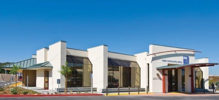 A modern, single-story medical clinic with large windows, a covered entrance, and a sign reading main community clinic on the facade, set against a clear blue sky.