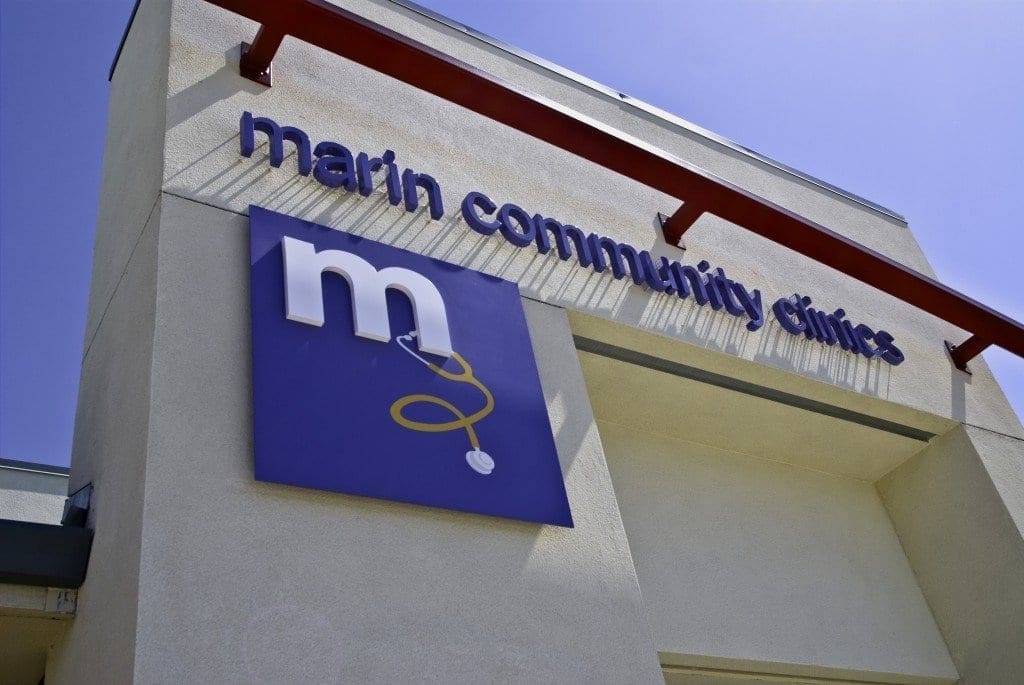 Exterior of a building with a sign that reads marin community clinics and a blue logo featuring a white m and a yellow stethoscope, viewed from below on a sunny day.