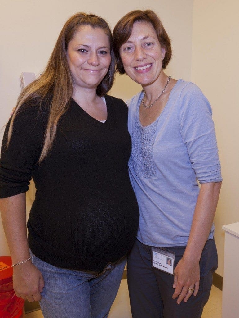 Two women standing together and smiling in a room; one appears to be pregnant and the other is wearing a name badge, suggesting she might be a healthcare worker.