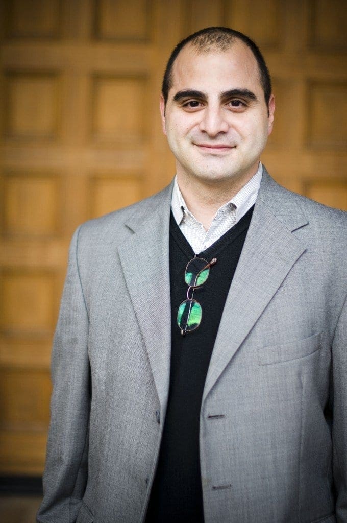 A man with short dark hair, wearing a gray suit jacket over a black sweater and collared shirt, stands in front of a wooden paneled background, smiling slightly. Glasses hang from the neckline of his sweater.