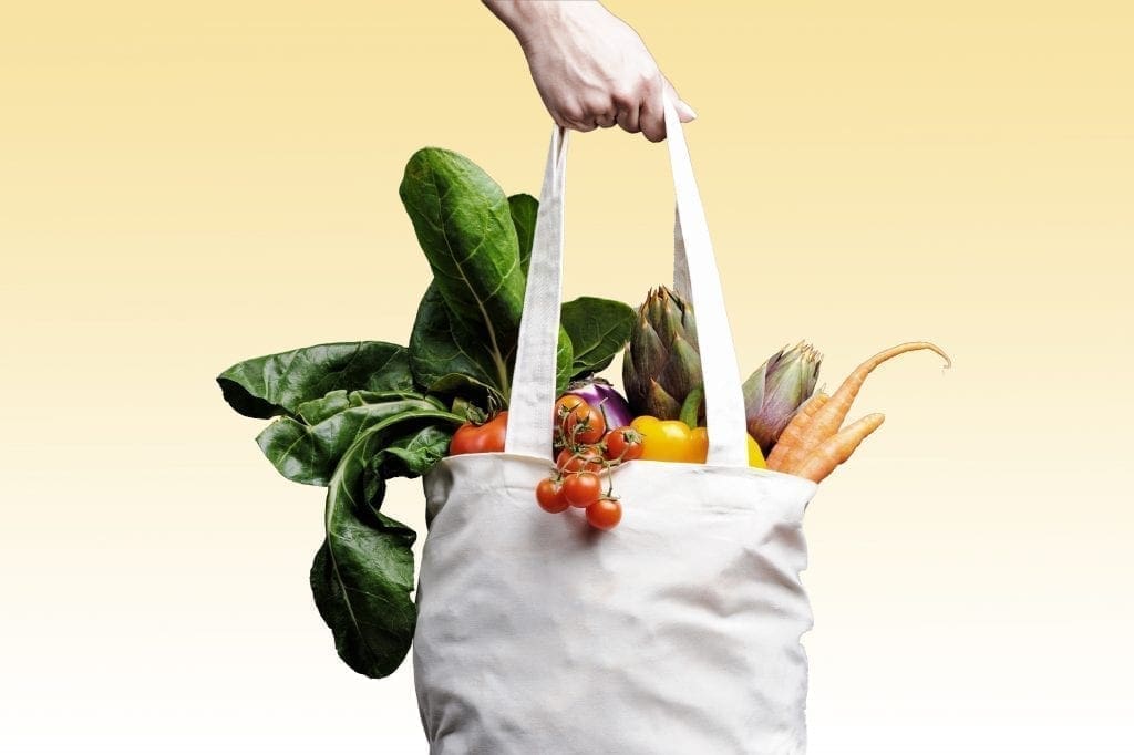A hand holds a white reusable tote bag filled with fresh vegetables, including carrots, artichokes, leafy greens, tomatoes, and cherry tomatoes, against a yellow gradient background.