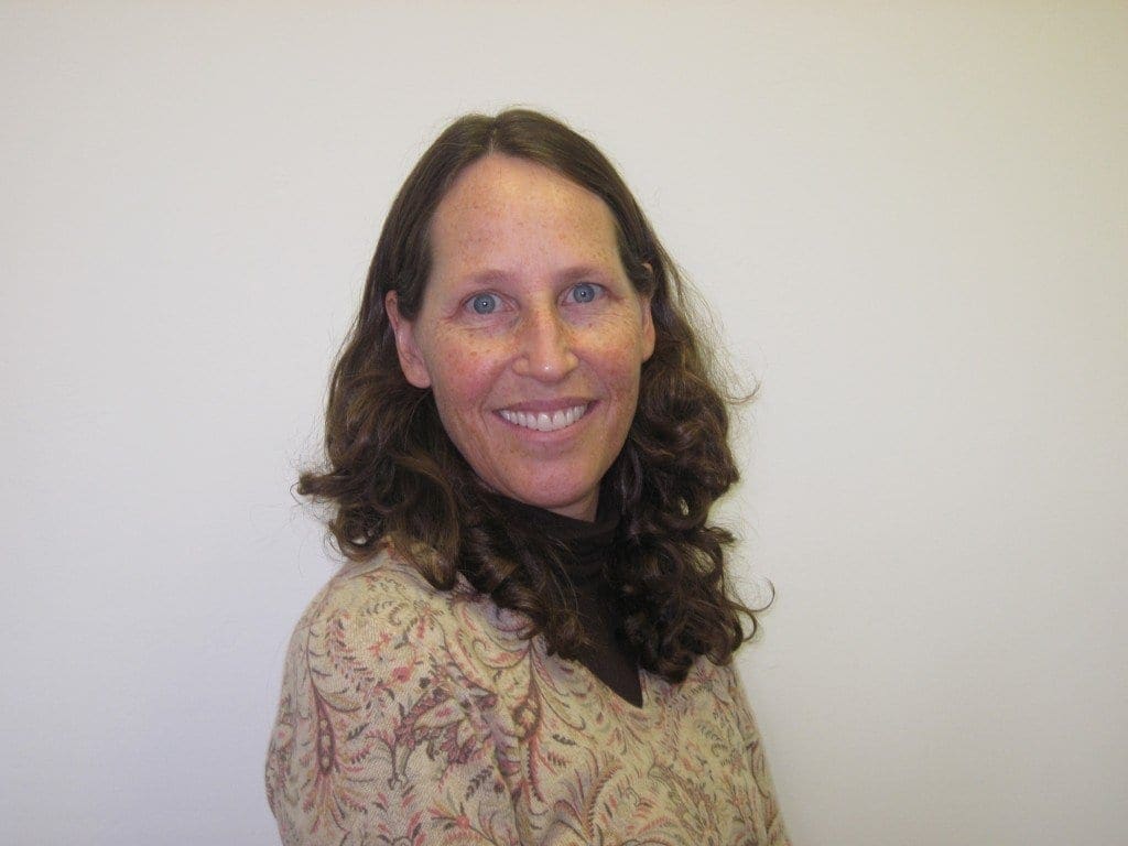 A woman with long wavy brown hair and blue eyes smiles at the camera. She is wearing a beige sweater with a floral pattern over a dark top, and stands in front of a plain light-colored background.