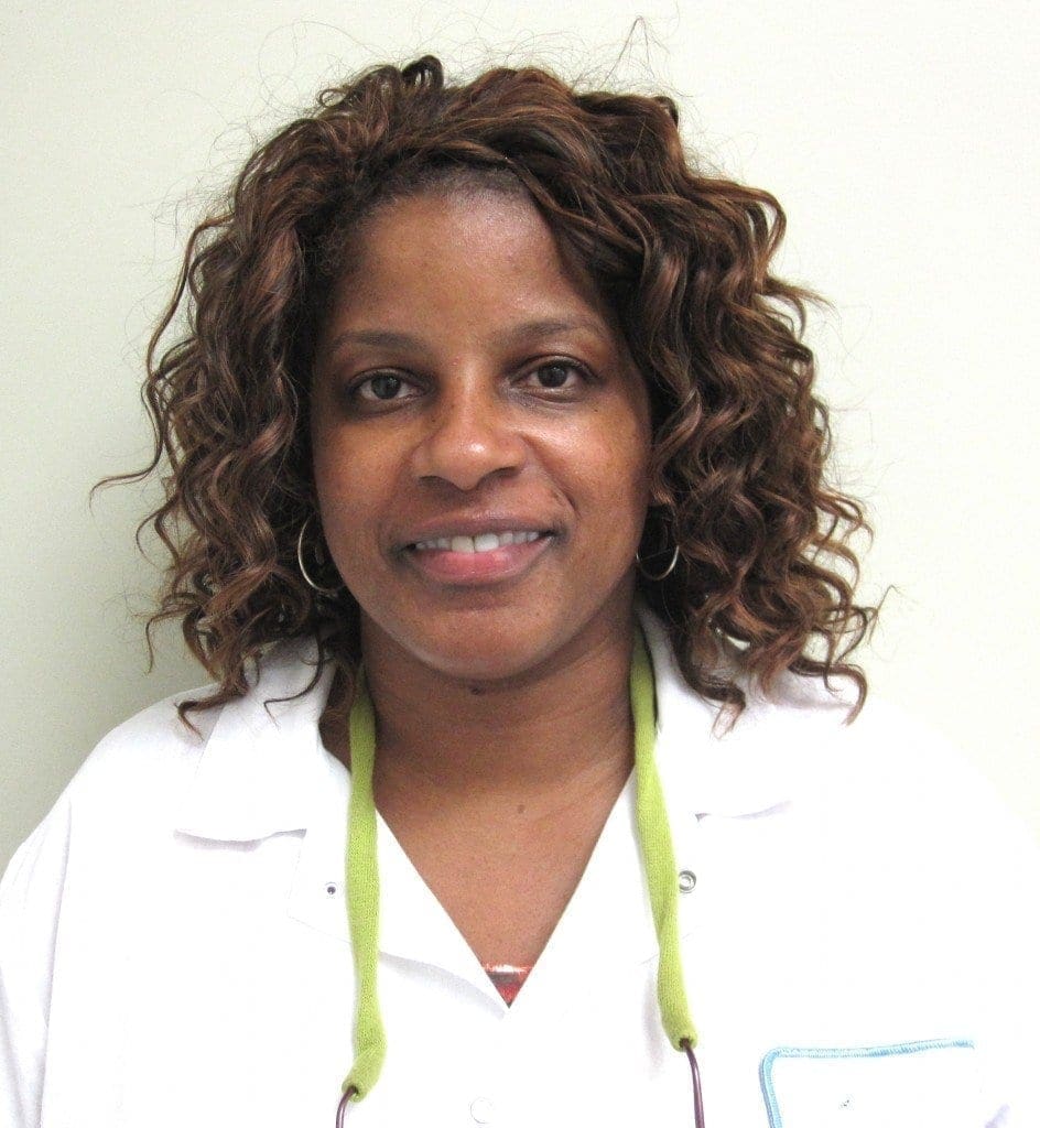 A woman with curly brown hair, wearing a white lab coat and a green lanyard, stands in front of a plain light-colored wall, smiling at the camera.