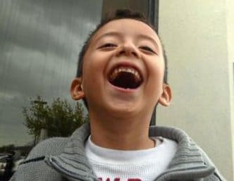 A young boy wearing a gray sweater laughs joyfully with his mouth wide open while standing outside near a window on a cloudy day.