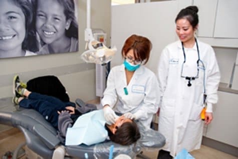 A child lies on a dental chair while a dentist examines their mouth, with another dental professional standing nearby. A black-and-white photo of smiling children hangs on the wall behind them.