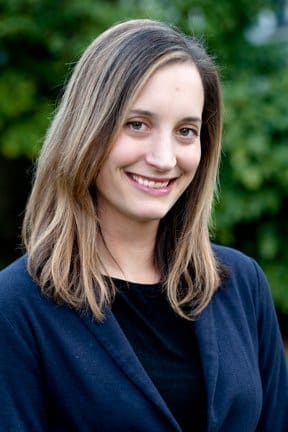 A woman with straight, shoulder-length brown hair smiles at the camera. She is wearing a dark blazer over a black top, and the background is blurred greenery.