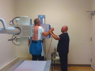 A man wearing a protective lead apron stands facing an X-ray machine as a technician adjusts his arm position in a medical examination room.