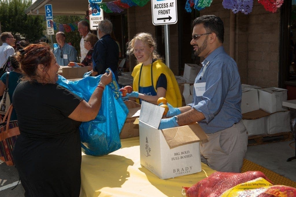 Volunteers stand behind a table outdoors, smiling as they hand out food items to a woman holding a large blue bag; boxes and people are visible in the background under colorful decorations.