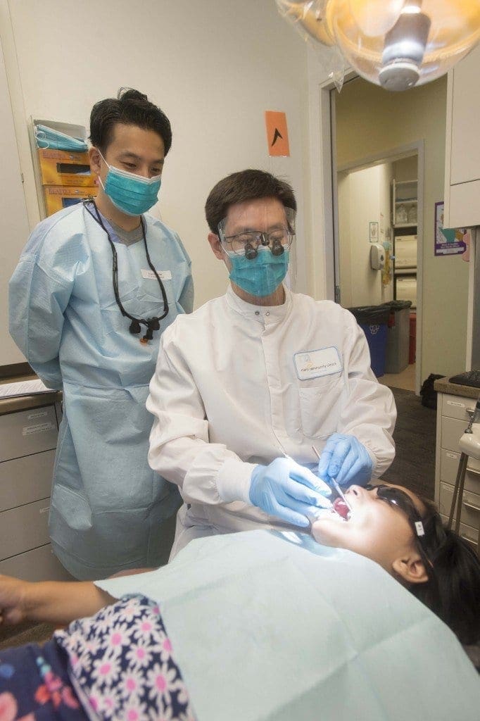 A dentist wearing a white coat and mask examines a young patient’s mouth while an assistant in blue scrubs and mask observes in a brightly lit dental office.