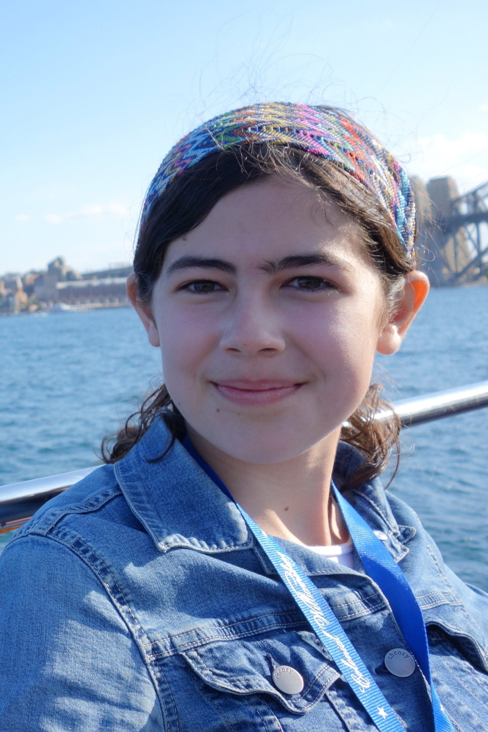 A young person with dark hair and a colorful headband smiles by the water, wearing a blue jacket and a lanyard, with a cityscape and bridge visible in the background.