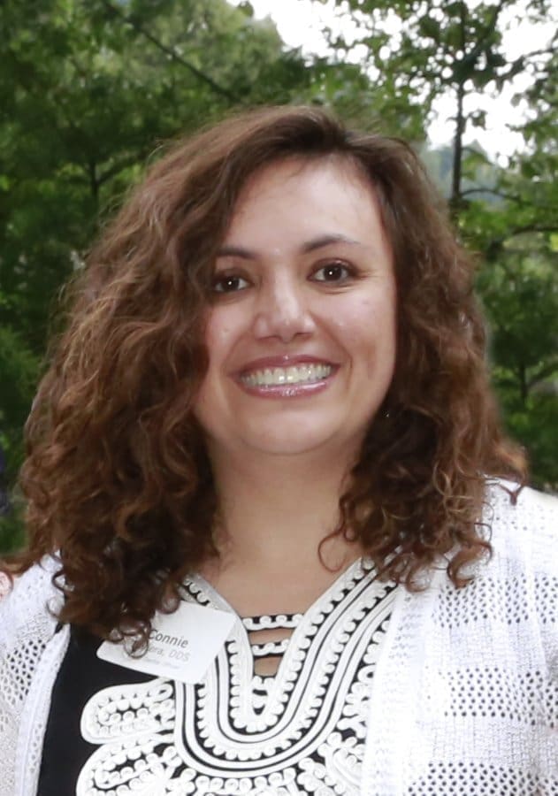 A woman with curly brown hair smiles at the camera. She is wearing a black and white patterned top and a white cardigan. There is greenery in the blurred background. She has a name tag on her chest.