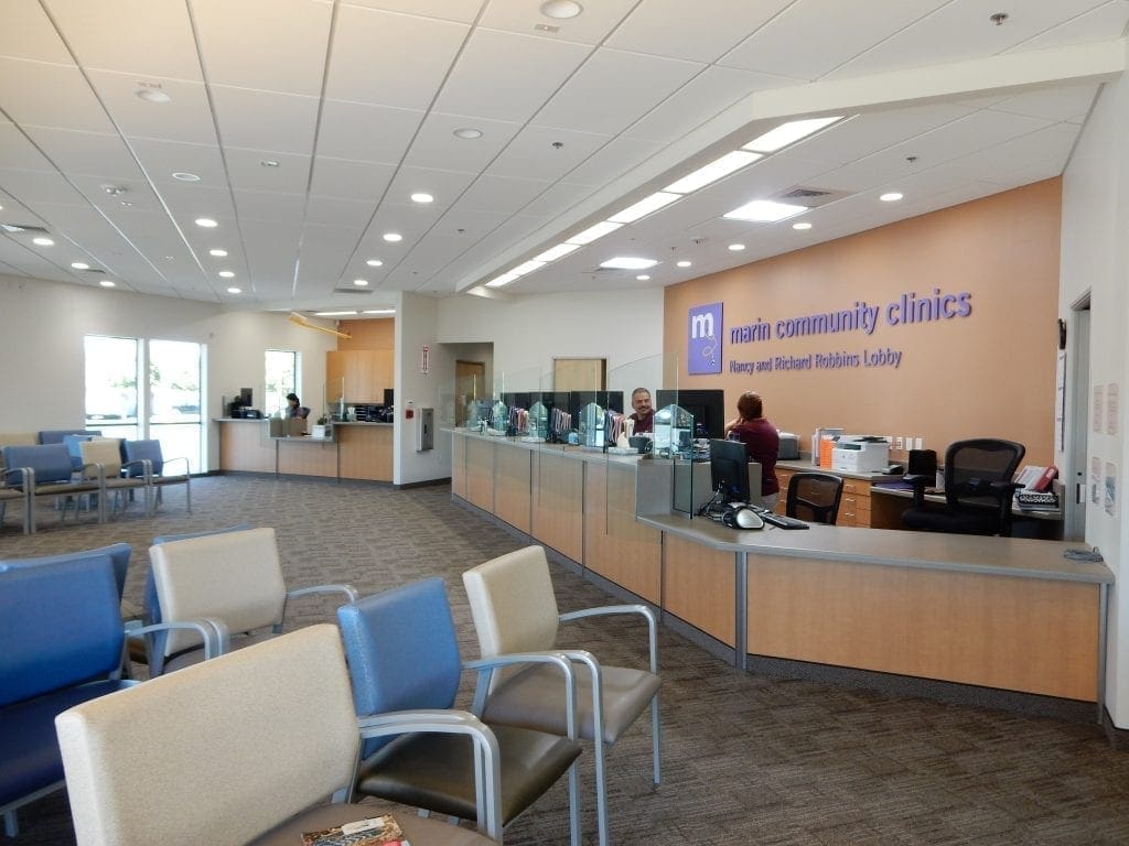 A modern medical clinic waiting area with rows of empty chairs and a front desk where staff assist patients. The wall behind the desk has a sign reading Marin Community Clinics.