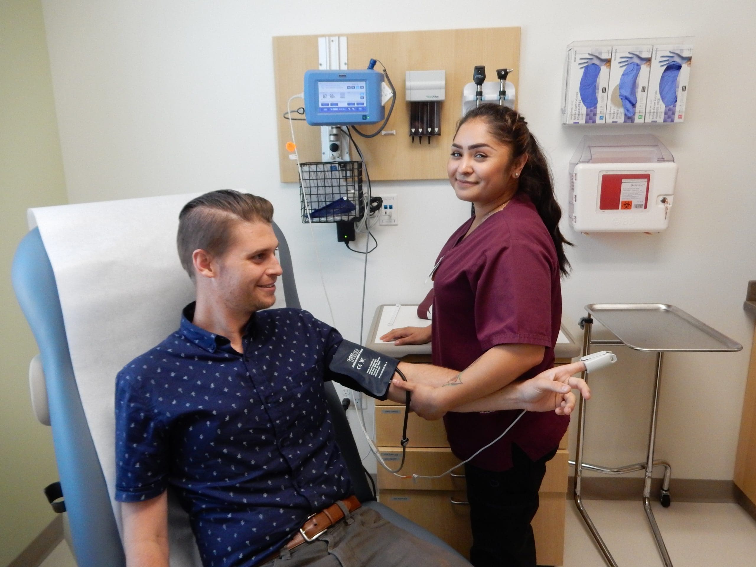 A nurse wearing scrubs checks a patients blood pressure in a medical exam room. The patient sits on an exam table, smiling, while the nurse stands beside him, holding the blood pressure monitor.