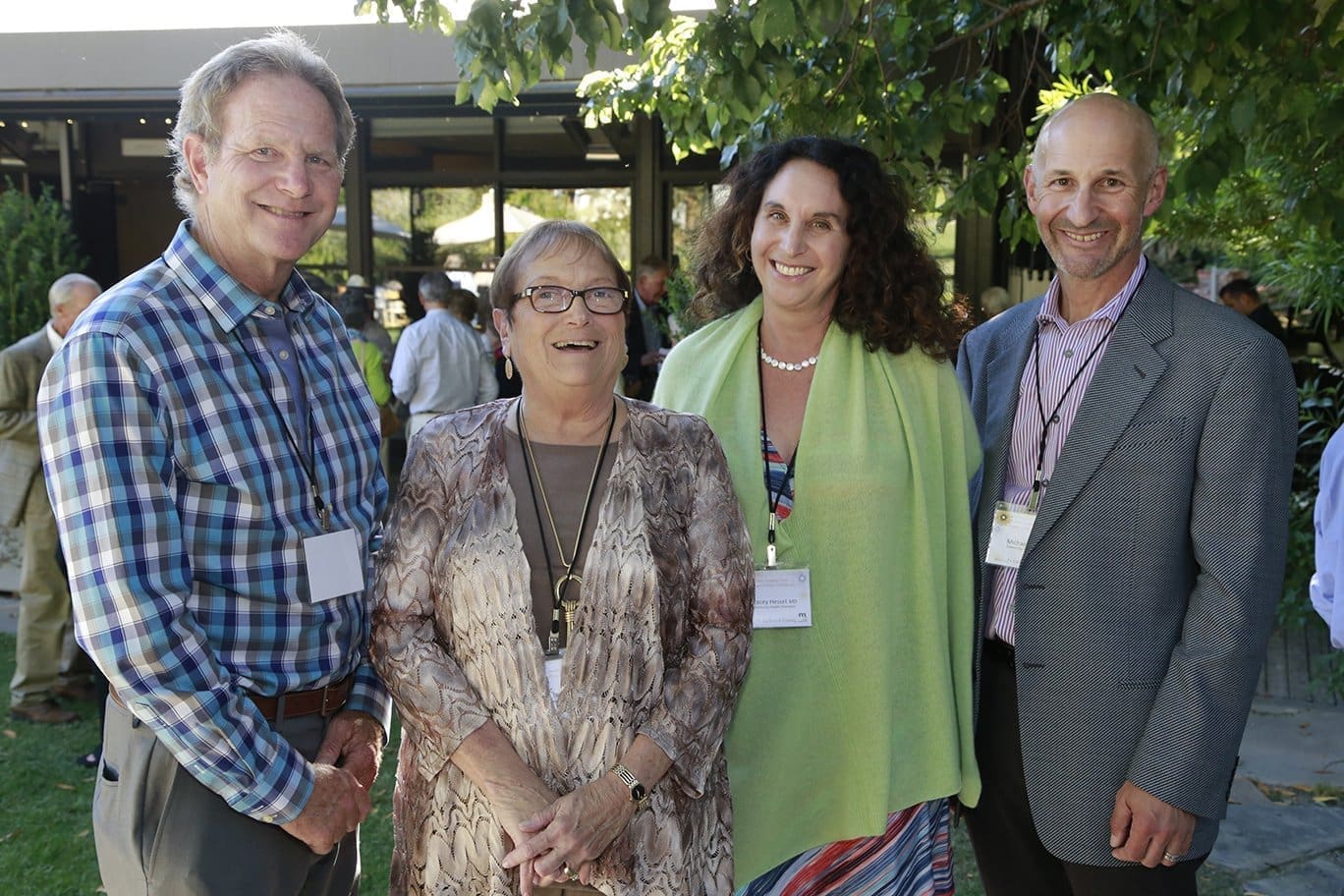 Four adults stand smiling outdoors at an event, all wearing name tags. Two men in dress shirts and blazers stand on either side of two women, one in a patterned dress and the other in a green shawl. Trees and people are in the background.