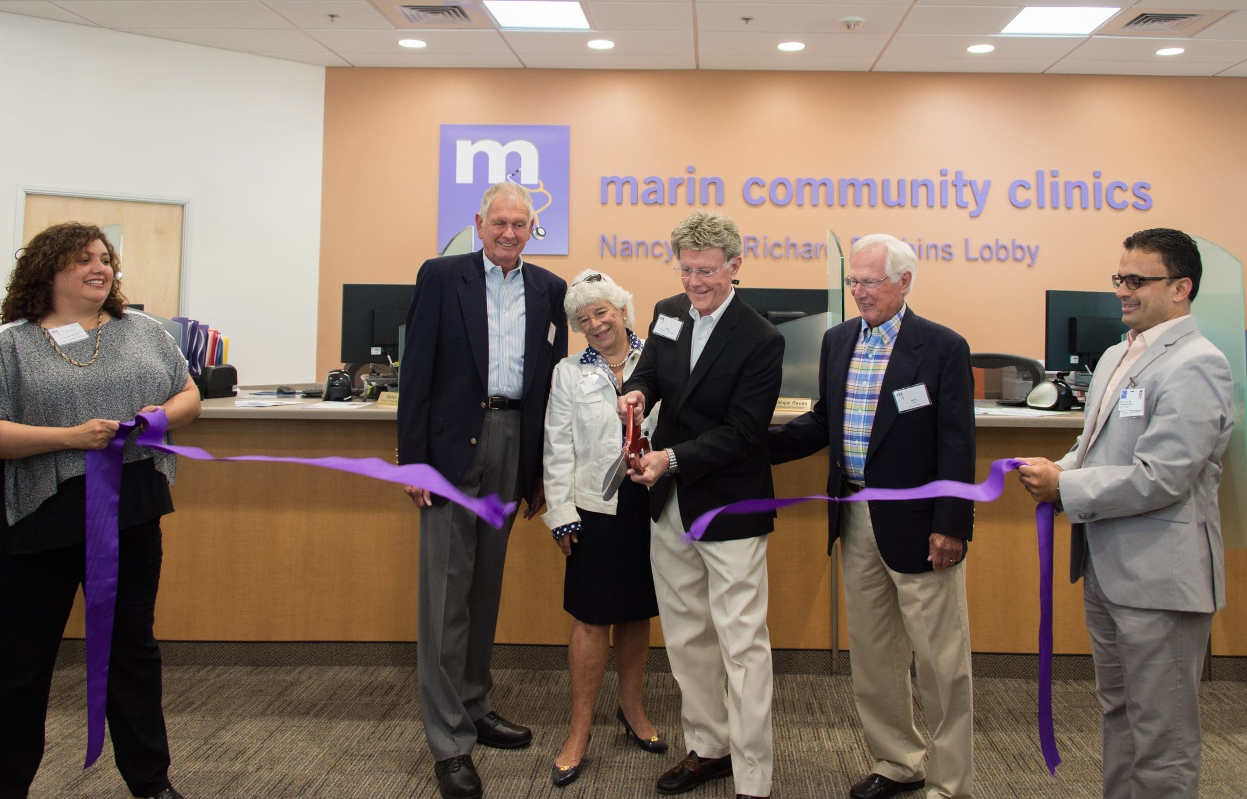 Six people stand in front of a reception desk at Marin Community Clinics, smiling as one person cuts a purple ribbon in a ribbon-cutting ceremony. A sign reads marin community clinics Nancy & Richard Robbins Lobby.