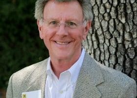 A smiling older man with gray hair and glasses, wearing a light gray blazer and white shirt, stands outdoors in front of a tree trunk.