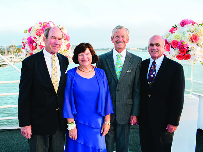 Four older adults, three men in suits and a woman in a blue dress, stand together smiling on a boat deck decorated with floral arrangements. Water and boats are visible in the background.