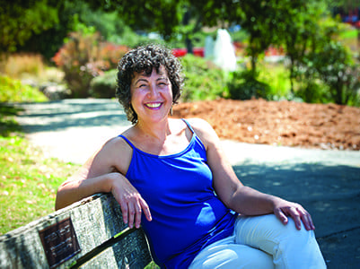 A woman with curly hair, wearing a blue sleeveless top and white pants, sits smiling on a wooden park bench on a sunny day, with greenery and a path in the background.
