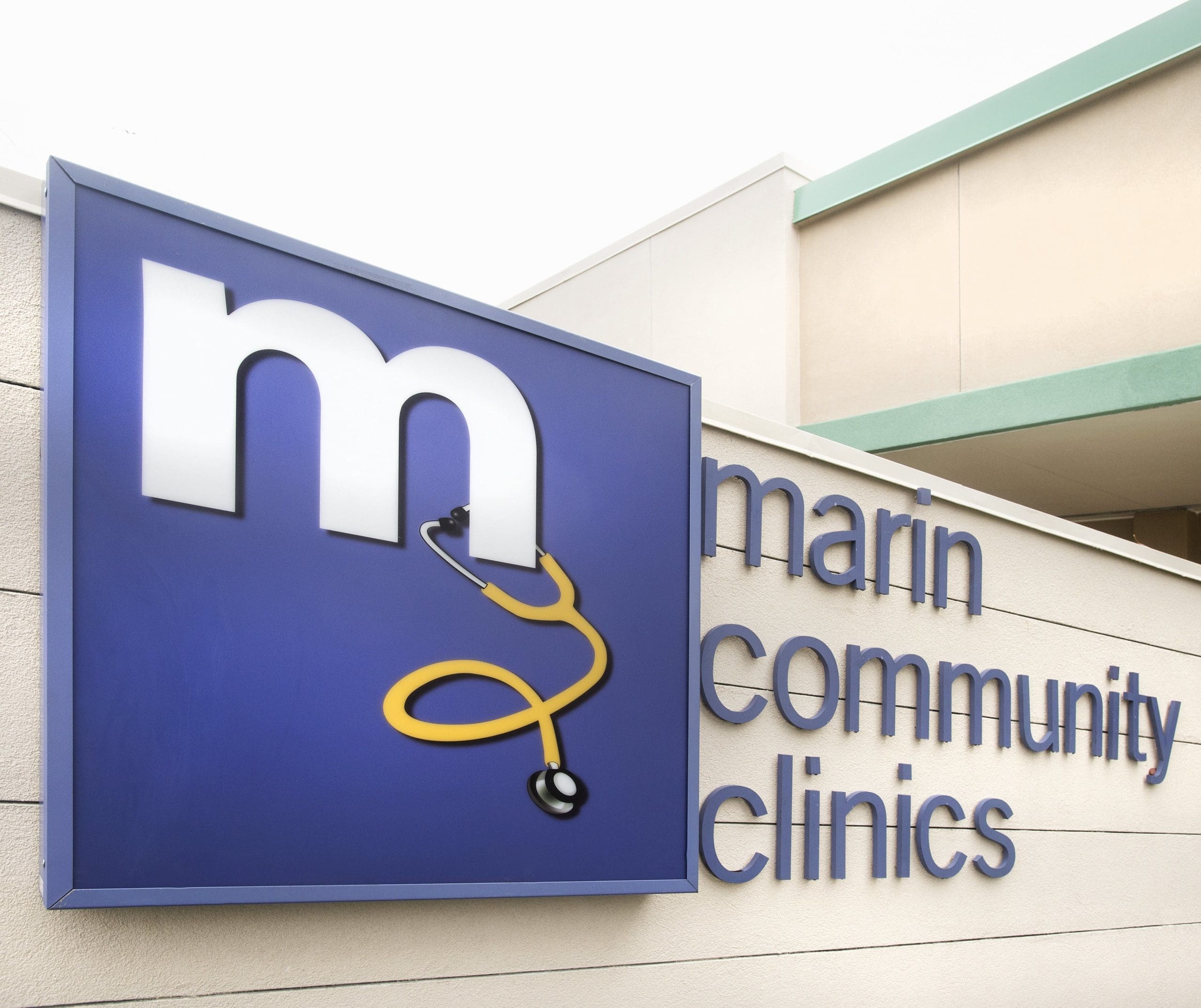 Large blue sign on a building reads marin community clinics with a white lowercase m and a yellow stethoscope forming part of the letter. The sign is mounted on a light-colored exterior wall.