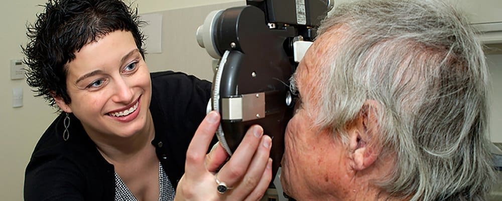 A woman smiles while examining an older mans eye using specialized ophthalmology equipment in a medical clinic.