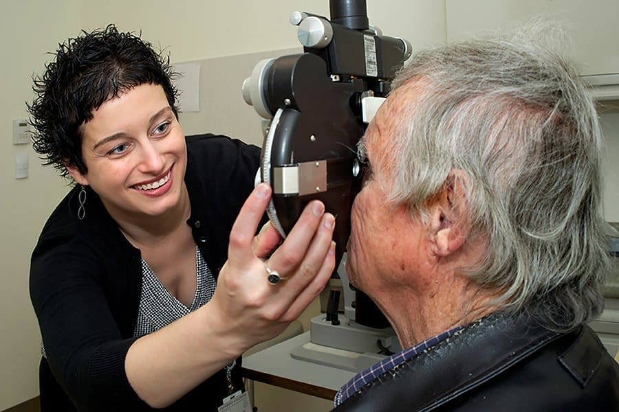 A smiling optometrist examines an older mans eyes using a slit lamp in a clinic setting. The man sits facing the device while the optometrist adjusts it.