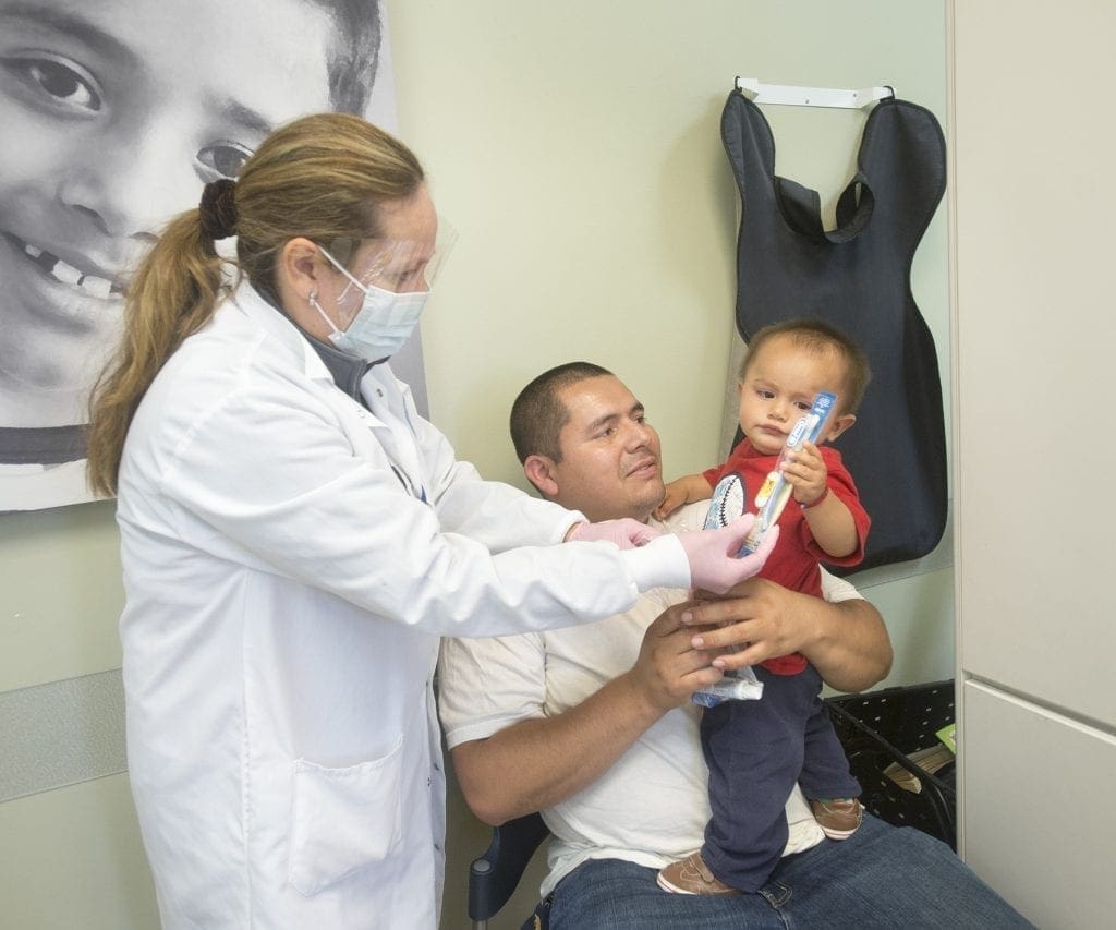 A dentist shows a young child how to use a toothbrush while the child sits on a mans lap. The dentist wears a mask and gloves. The child holds a toothbrush and a tube. A dental X-ray apron hangs on the wall behind them.