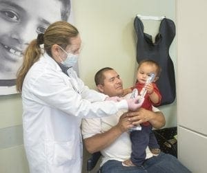 A dentist shows a young child how to use a toothbrush while the child sits on a mans lap. The dentist wears a mask and gloves. The child holds a toothbrush and a tube. A dental X-ray apron hangs on the wall behind them.