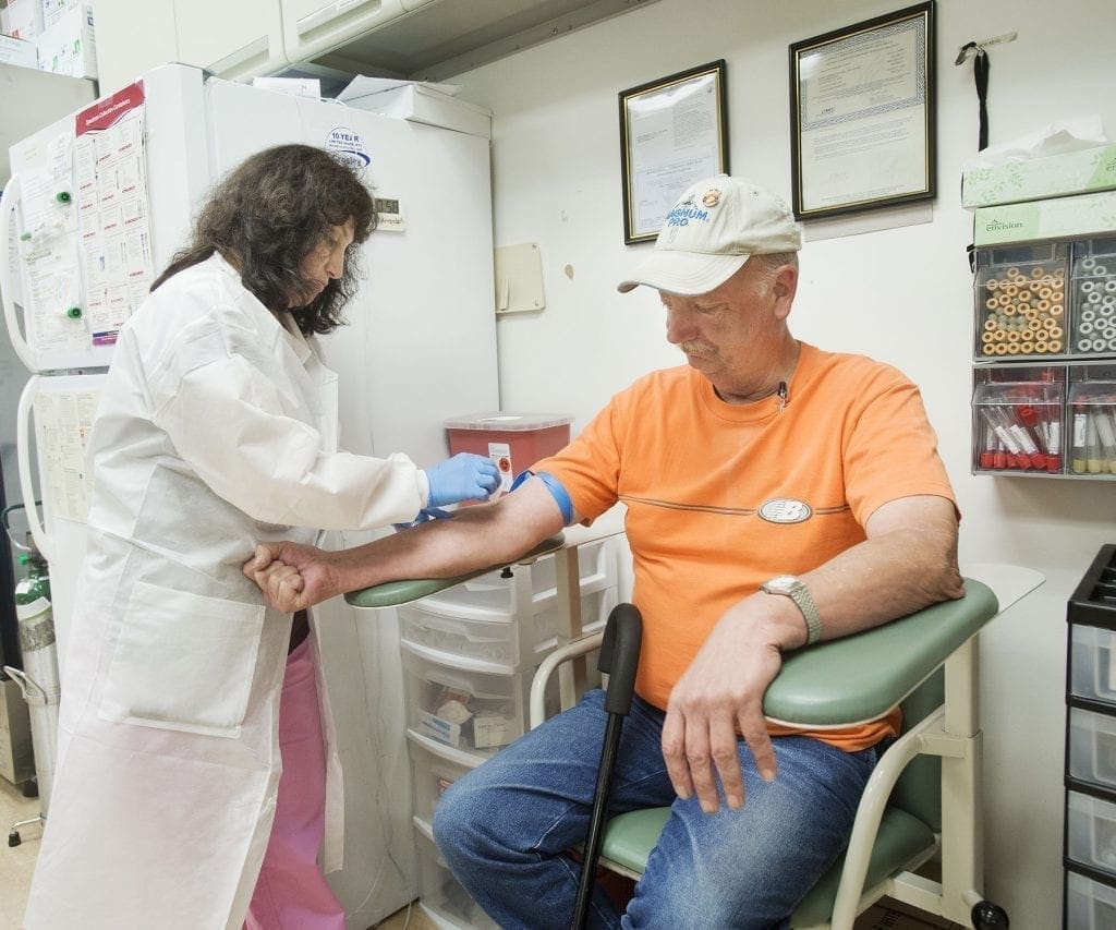 A healthcare worker in a white lab coat draws blood from a seated man wearing an orange shirt and blue jeans in a medical office. The man rests his arm on a green armrest while the worker prepares his arm with a tourniquet.