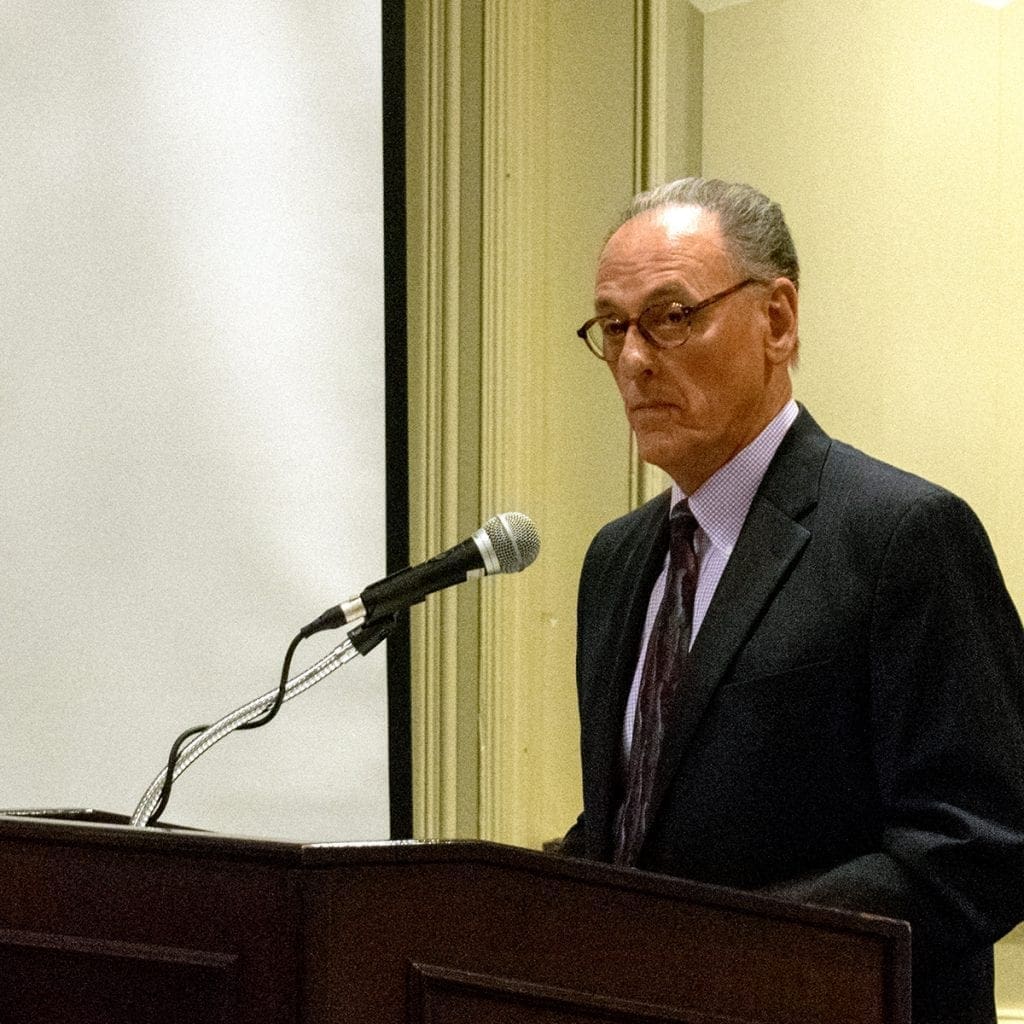 A man in a suit and tie stands at a podium with a microphone, facing forward. He is indoors, next to a large white screen and beige walls, appearing to speak or present at an event.