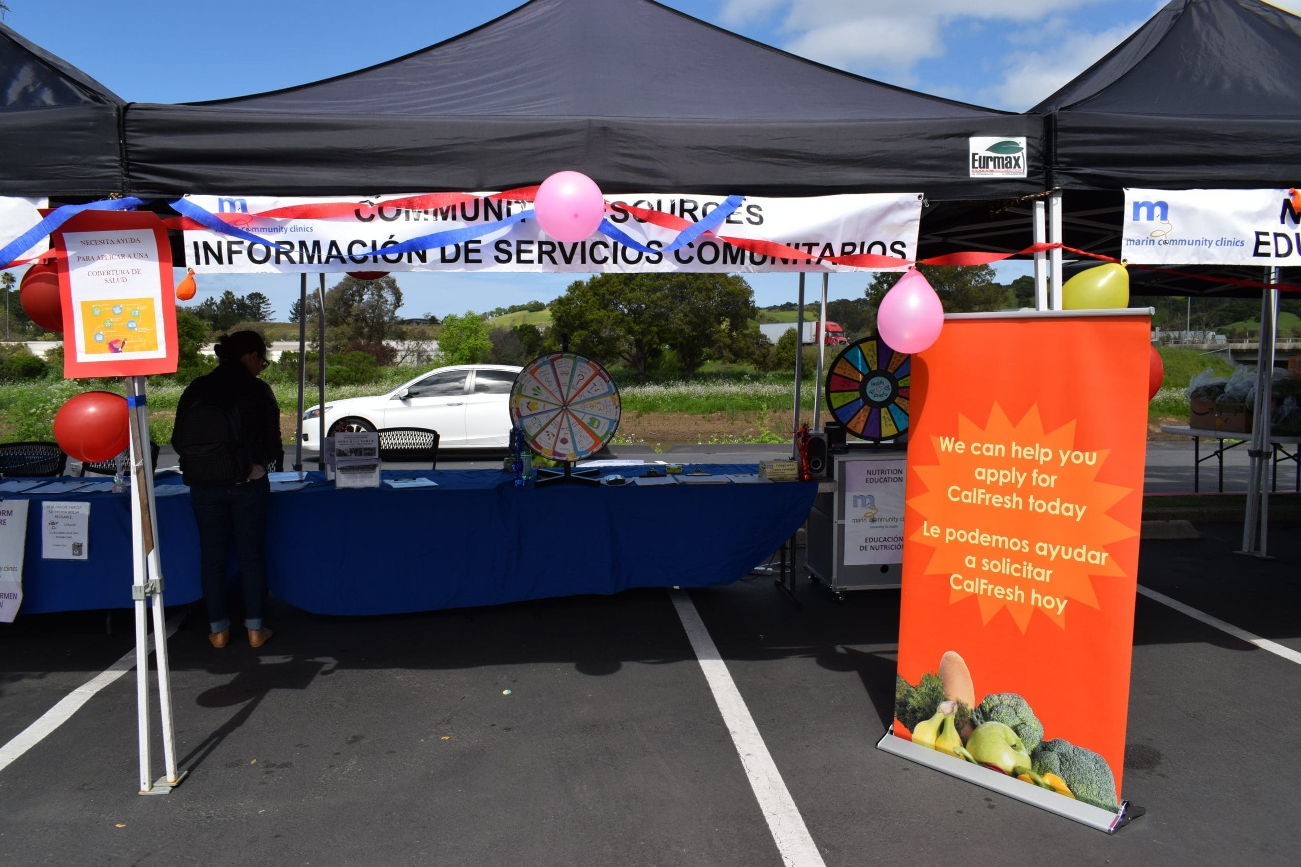 A community event booth with informational banners, colorful balloons, two prize wheels, and a sign offering help to apply for CalFresh in English and Spanish. The booth is outdoors under a tent.