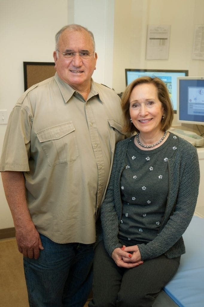 A man in a beige short-sleeve shirt stands next to a seated woman in a gray sweater and patterned shirt. They both smile at the camera in a room with medical equipment and computer monitors in the background.